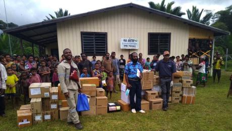 all unloaded medical supplies' boxes nicely stacked in front of the Haia airstrip building, with members of the community in the back and the health officer and one of the PI officers in front