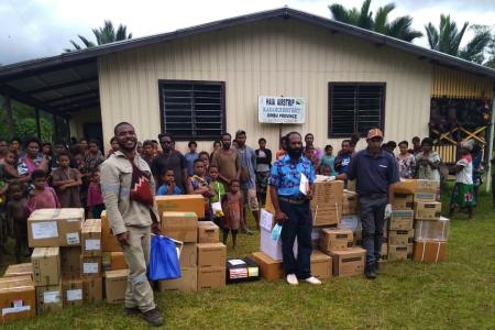 all unloaded medical supplies' boxes nicely stacked in front of the Haia airstrip building, with members of the community in the back and the health officer and one of the PI officers in front