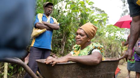 A Kabwum woman sits in a wheelbarrow as her husband and son explain her condition.